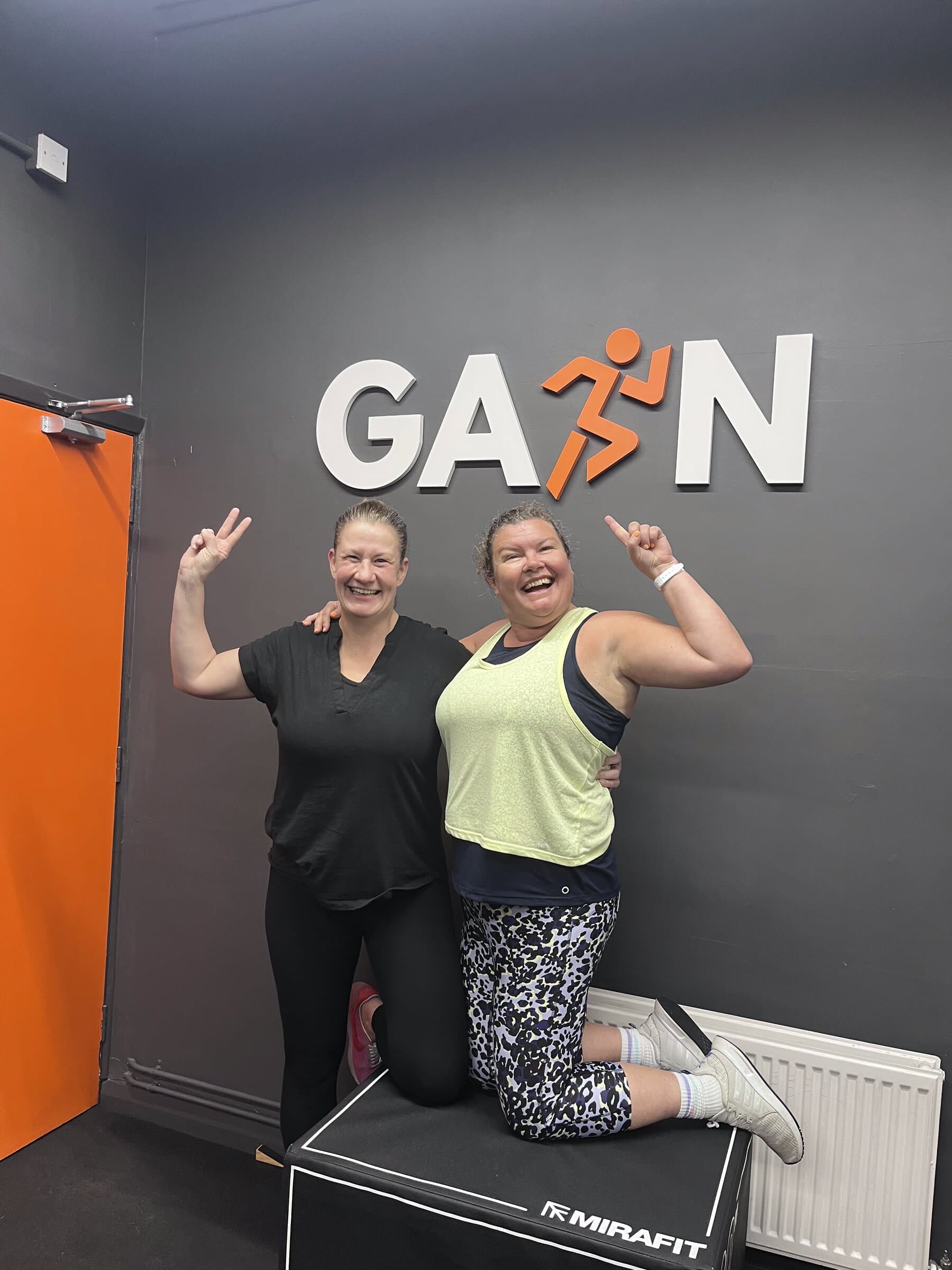 Two women smiling in front of the GAIN sign in the gym